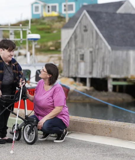 Person using a walker and holding a white cane stands on a waterfront path while another person kneels beside them, speaking with them; a dock, boat, and weathered buildings are visible in the background.