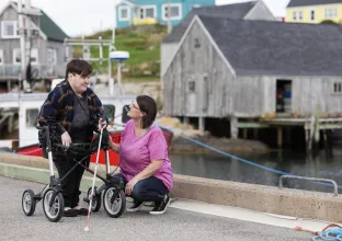 Person using a walker and holding a white cane stands on a waterfront path while another person kneels beside them, speaking with them; a dock, boat, and weathered buildings are visible in the background.