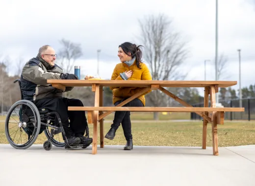 Two people sitting at a picnic table outside. Both are smiling. One person is in a wheelchair. 
