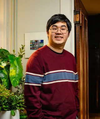 Young man standing in front of plants with his hands in his pockets, smiling.