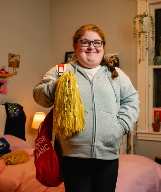 Smiling young lady, standing in a bedroom, holding a backpack and cheerleading pom pom. 