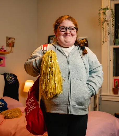 Smiling young lady, standing in a bedroom, holding a backpack and cheerleading pom pom. 